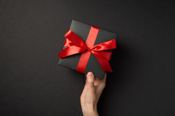 First person top view photo of woman hand holding stylish black giftbox with red ribbon bow on isolated black background