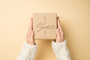 First person top view photo of girl hands in white knitted pullover holding craft paper giftbox with twine bow on isolated beige background