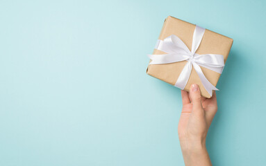 First person top view photo of hand holding stylish craft paper giftbox with white satin ribbon bow on isolated pastel blue background with copyspace