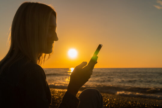 Close up young woman is sitting on a pebble beach by the sea with a mobile phone in her hands at sunset, a photo against the sun. Concept of happiness, travel, lifestyle