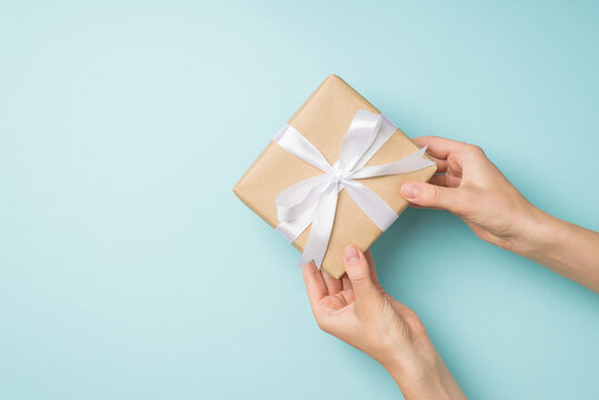 First person top view photo of hands giving craft paper giftbox with white satin ribbon bow on isolated pastel blue background with copyspace