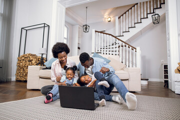 African american family of four sitting together on floor and watching cartoon on laptop. Young parents with two daughters using technology during free time at home.