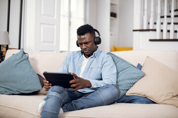 Young african man in wireless headphones resting on comfy couch with digital tablet in hands. Handsome guy in casual clothes surfing internet or listening to music during free time at home.