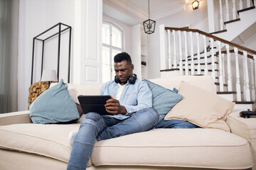 Fototapeta premium Young african man in wireless headphones resting on comfy couch with digital tablet in hands. Handsome guy in casual clothes surfing internet during free time at home.