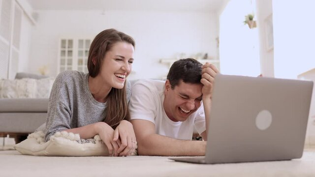 Loving couple using laptop and laughing while lying on floor in apartment room spbi. Closeup view of young woman, man look at computer screen and laugh, talk with smiles and lie in light interior. Two
