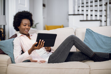 Obraz premium Pretty smiling african american woman lying on cozy sofa and watching movie on digital tablet. Pleasant female enjoying free time at home with modern device.
