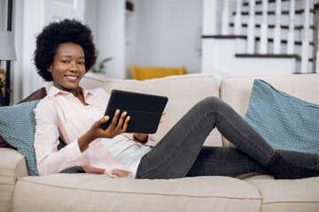 Pretty african woman smiling on camera while lying on couch with digital tablet in hands. Young female in casual wear reading news in internet during free time.