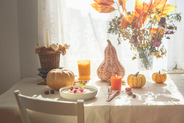 Autumn breakfast table with pumpkins, bunch of orange fall leaves and flowers, waffles with whipped cream and candles. Domestic still life at window with sunlight