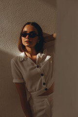 Stylish woman in white outfit and in retro sunglasses standing near white wall on sunny day in city and looking at camera 