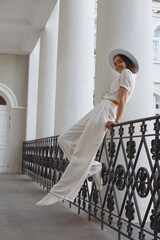 Side view of coquettish bride in fancy white outfit and hat leaning on fence in city and looking at...