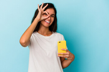 Young caucasian woman holding a mobile phone isolated on blue background excited keeping ok gesture on eye.