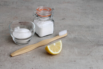 Baking soda in a glass jar, lemon and bamboo toothbrush on a gray concrete background with copy space. Natural teeth whitening at home. 