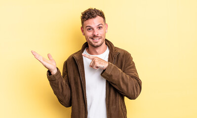 young handsome hispanic man smiling cheerfully and pointing to copy space on palm on the side,...