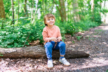 Little preschool boy sitting on a tree trunk in a forest and playing with wooden stick. Child spending time outdoor in summer. Toddler in a park.