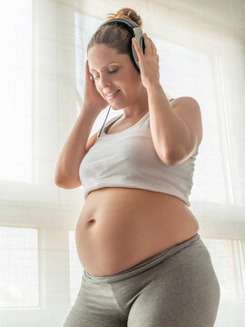 happy pregnant woman listening to music on headphones with a light and a window in the background.