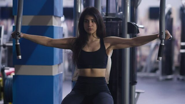 Portrait of confident concentrated Middle Eastern woman exercising in seated chest fly machine. Front view medium shot of fit slim beautiful sportswoman training muscles with equipment in gym indoors