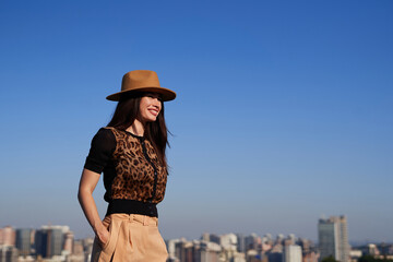 Shopping time concept: attractive fashion styled brunette woman in leopard pattern blouse, trousers and brown hat on the background of clear blue sky and urban background with space for text or ads