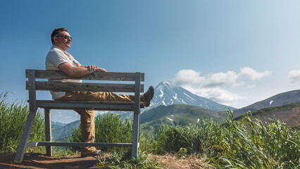 Naklejka na ściany i meble A man in sunglasses is sitting on a bench and smiling. One leg is lying on the seat. There is green grass around. There is a conical volcano with snow-covered slopes against a blue sky. Kamchatka