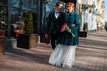 Naklejka premium the bride and groom are walking along the city street in wedding clothes and with a bouquet of tulips