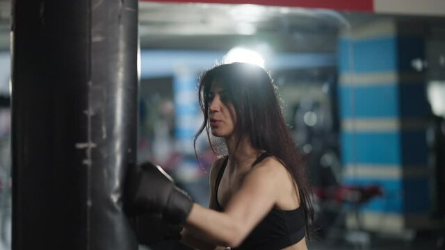 Live camera follows movement of confident motivated female boxer punching bag in gym light. Middle Eastern fit slim beautiful sportswoman training indoors. Martial arts and self-defense concept