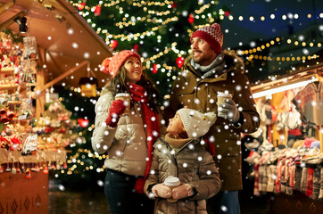 family, winter holidays and celebration concept - happy mother, father and little daughter with takeaway drinks at christmas market on town hall square in tallinn, estonia over snow