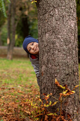 Naklejka na ściany i meble Cute preschool boy peeking out from behind a tree in the park. Boy smiling and looking at the camera