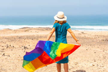 A lesbian person with a green dress, a white hat and with the LGBT flag by the sea, a symbol of homosexuality