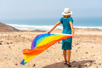 A lesbian person with a green dress, a white hat and with the LGBT flag by the sea, a symbol of homosexuality