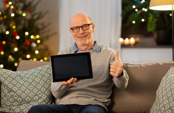 technology, winter holidays and people concept - happy smiling bald senior man with tablet pc computer sitting on sofa and showing thumbs up at home in evening over christmas tree lights background