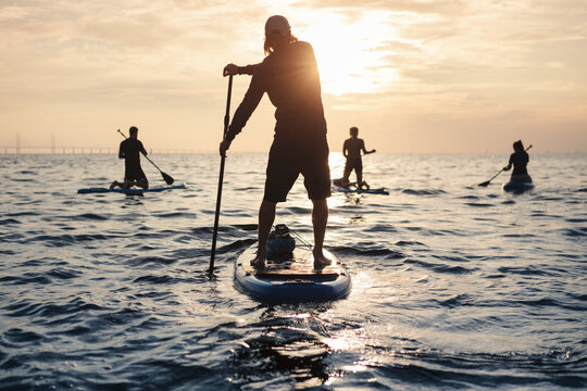 Male and female friends rowing paddleboard in sea during sunset