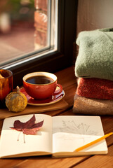 season and objects concept - open sketchbook with drawing of autumn leaf, cup of coffee, wool sweaters and pumpkin on wooden window sill at home