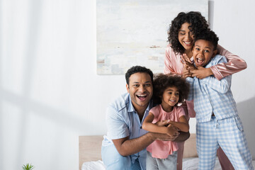 Fototapeta na wymiar happy african american parents hugging kids while having fun in bedroom in morning