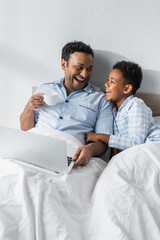 laughing african american father and son looking at each other near laptop in bed