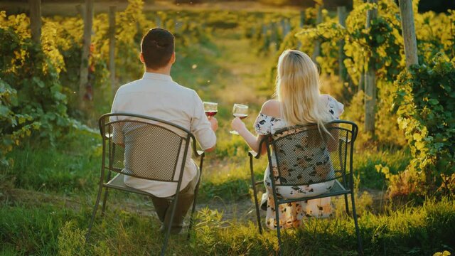 A Young Multi-ethnic Couple Is Relaxing By The Vineyard, Drinking Wine And Watching The Sunset. Wine Tour