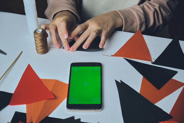 Children's hands make a homemade garland of orange and black triangles there is a phone with a green screen on the table