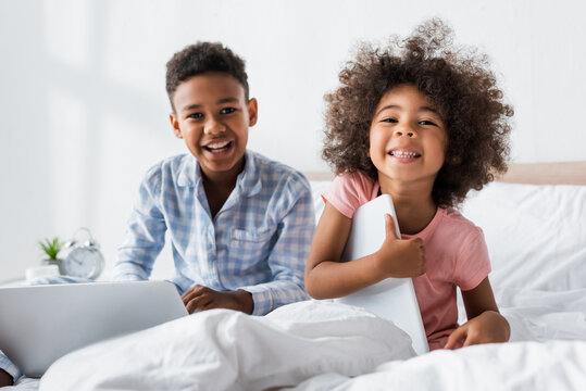 excited african american kids looking at camera while sitting with gadgets in bed