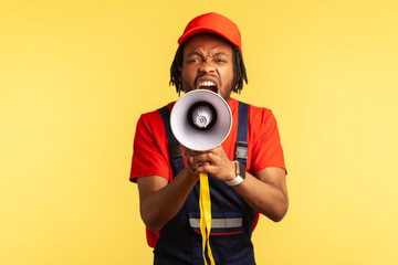 Portrait of angry bearded worker wearing red T-shirt and blue overalls holding megaphone and screaming with aggressive expression, protesting. Indoor studio shot isolated on yellow background.