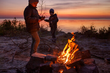 Kids of different ages sit near fire on autumn seashore after sunset, communicate and have a good...