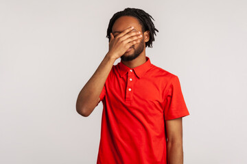 Man with dreadlocks wearing red casual style T-shirt, closing eyes with hand, dont want to see that, ignoring problems, hiding from stressful situations. Indoor studio shot isolated on gray background