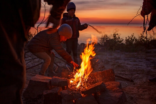 Kids Of Different Ages Sit Near Fire On Autumn Seashore After Sunset, Communicate And Have A Good Time Together, Trust And Friendship, Active Family Weekend. Autumn Outside Lifestyle. Focus On Fire