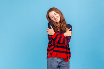Portrait of lovely female in striped casual style sweater, standing hugging herself and feeling good, appreciating her appearance, self esteem concept. Indoor studio shot isolated on blue background.