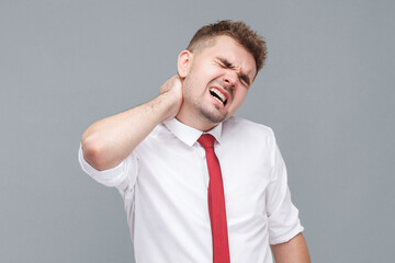 Neck ache. Portrait of young sick man in white shirt and tie standing and touching or massaging his painful neck. indoor isolated on gray background.