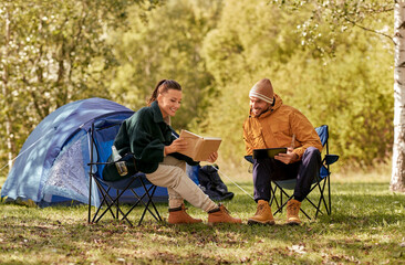 camping, tourism and travel concept - happy couple with tablet pc computer and book resting at tent camp