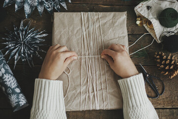 Hands wrapping stylish christmas gift in craft paper with string on rustic wooden table with blue paper stars. Flat lay. Atmospheric moody image, nordic style. Merry Christmas and Happy Holidays!