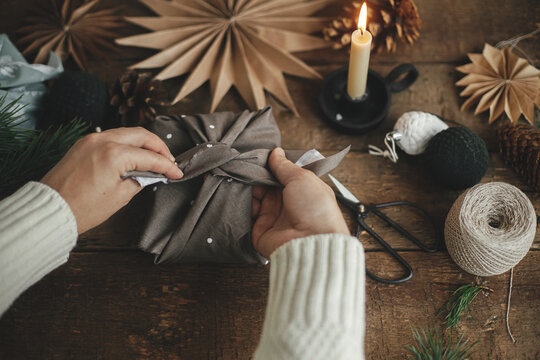 Furoshiki Christmas gift. Hands wrapping christmas gift in brown fabric on rustic wooden table with scissors, craft paper star, candle. Atmospheric moody time, nordic style. Zero waste holiday