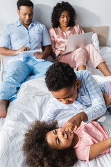 Obraz premium cheerful african american siblings having fun in bed near blurred parents with gadgets