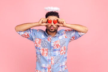 Romantic angelic man with beard covering eyes with small red hearts, making pout lips, kissing...