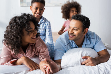 happy african american man holding hand of wife near blurred kids in bedroom
