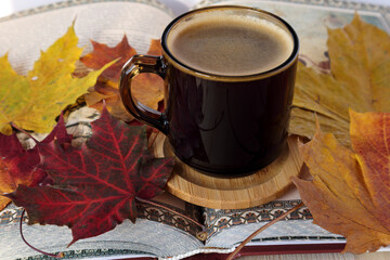 a cup of coffee on a wooden stand surrounded by autumn maple leaves on a centerfold book in sunlight - obrazy, fototapety, plakaty