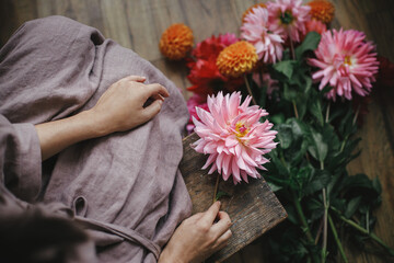 Woman holding pink dahlia flower and sitting on wooden rustic bench, view above. Atmospheric moody image. Florist in linen dress arranging autumn flowers bouquet in countryside. Slow life
